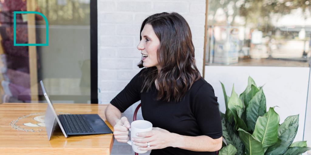 Photo of Jesica D'Avanza, a white woman with long brown hair wearing a black dress and holding a white coffee mug, pointed toward the left talking to someone out of view with a computer on the table
